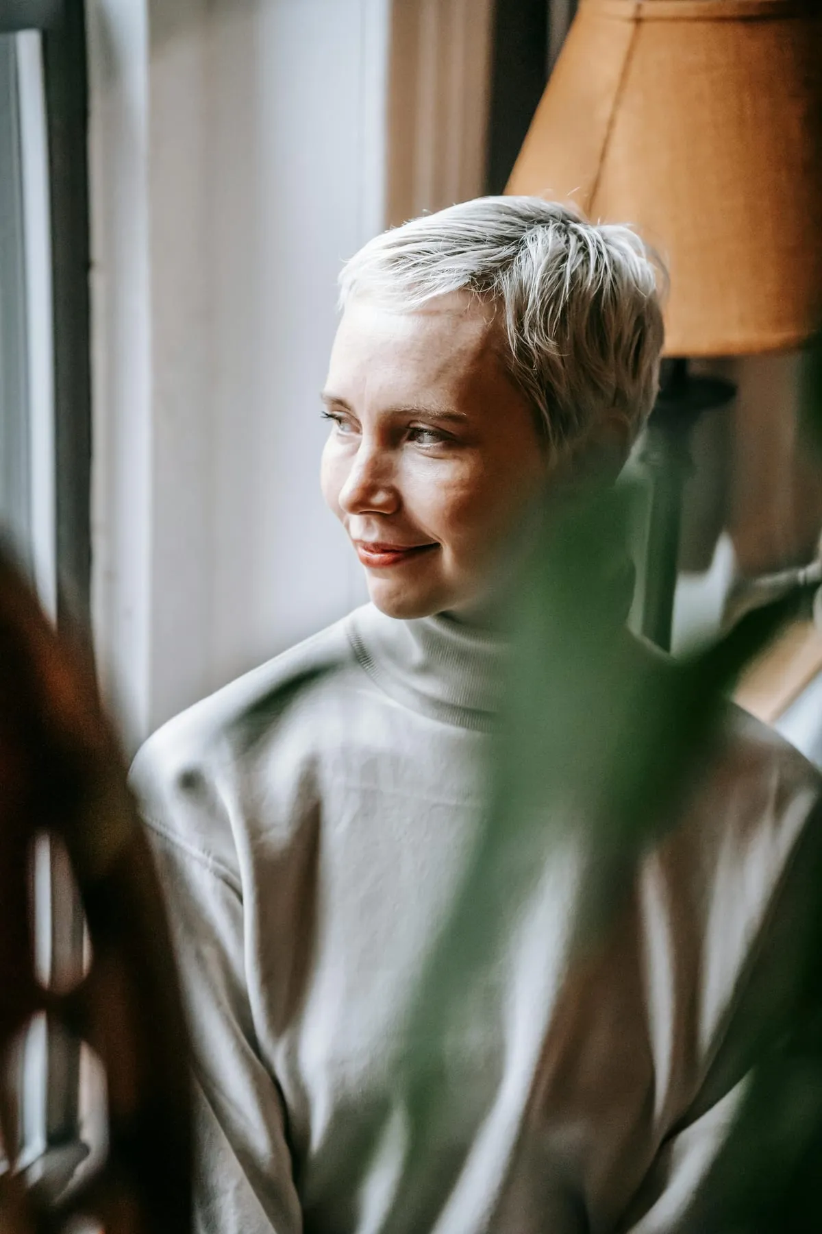 A woman with short grey hair looking out of a window, half-framed by a houseplant